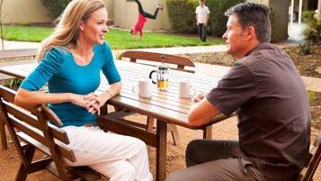 Zwei Personen sitzen an einem Tisch im Freien mit einer Kaffeekanne, in einem Innenhof mit einem Gebäude im Hintergrund.
