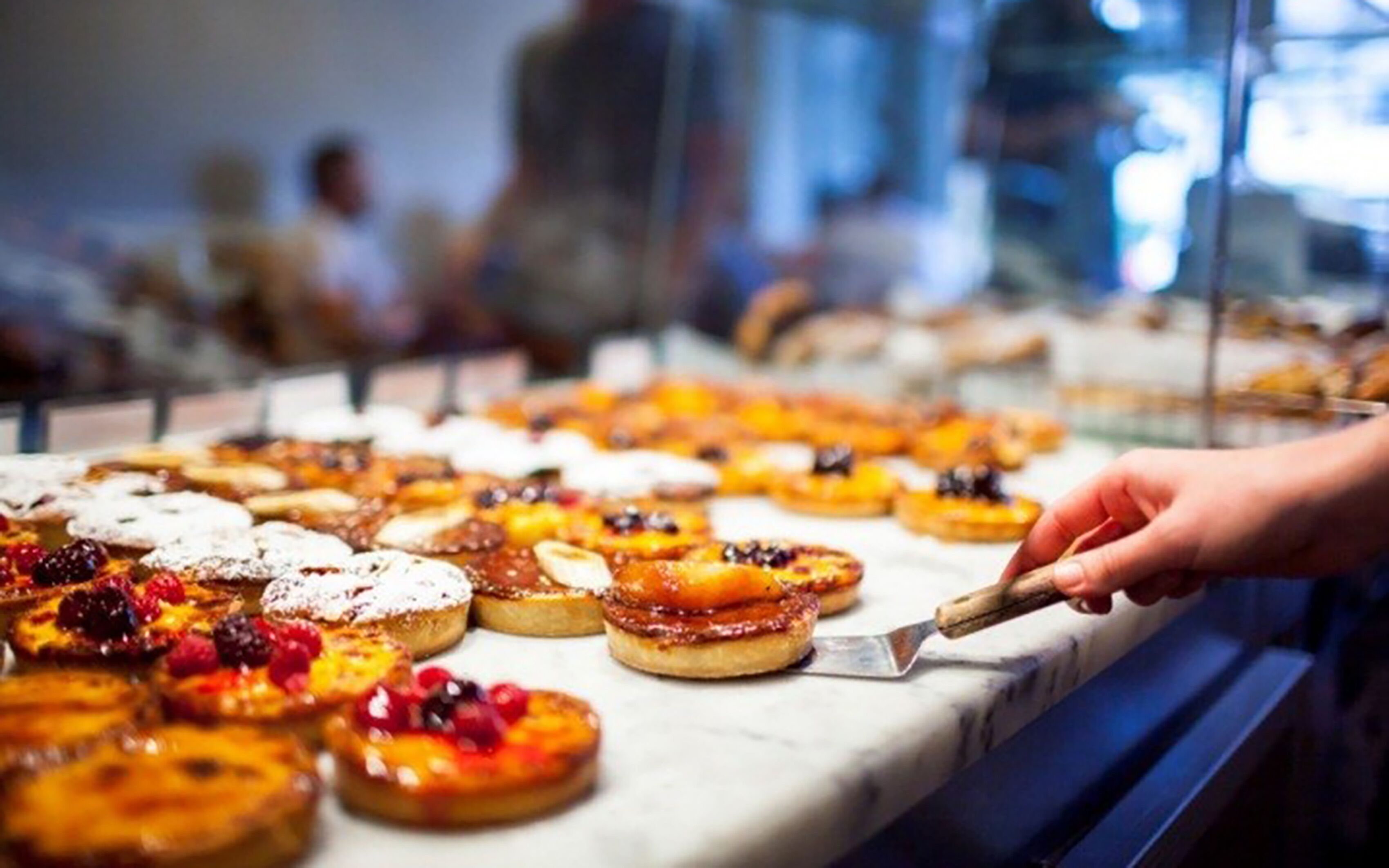 Diverse gebakjes in etalage, waaronder vruchten-toegesneden taartjes en desserts, terwijl een hand er een pakt.
