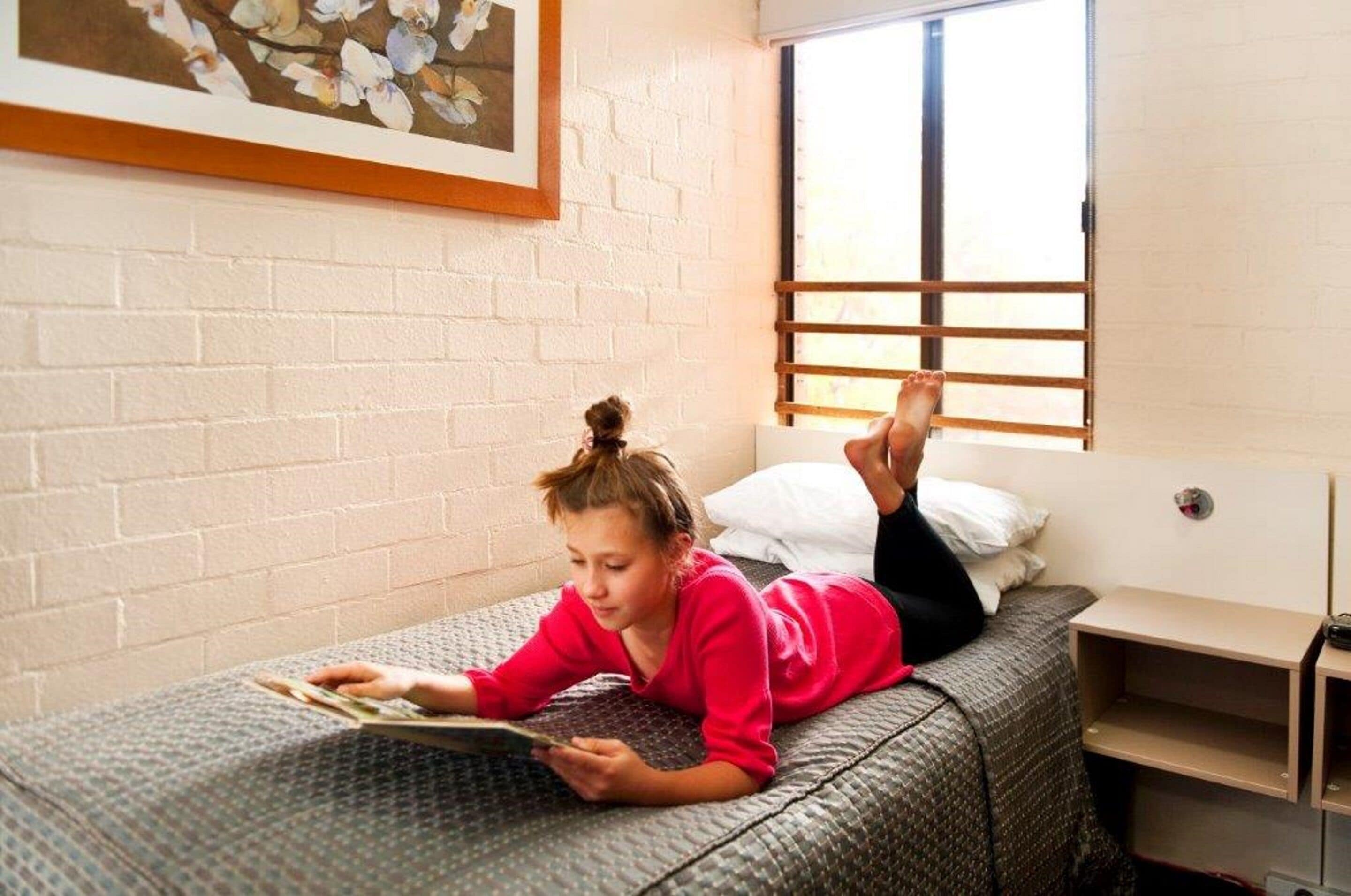 A room with a bed, a window with blinds, a small bedside table, and a wall-mounted shelf.
