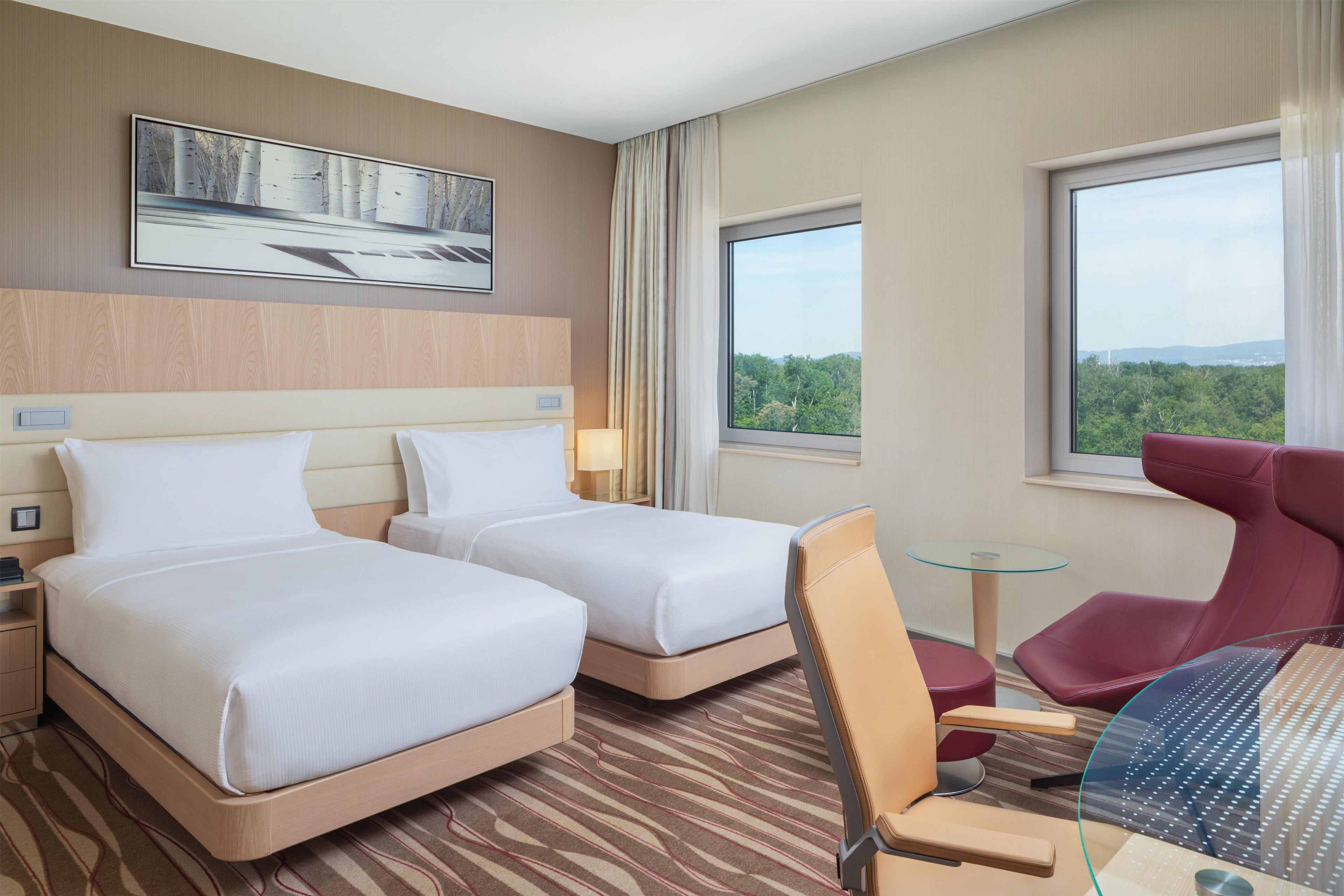 Une chambre d’hôtel avec deux lits, un bureau, une chaise, une petite table et une grande fenêtre donnant sur l’extérieur.