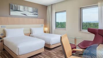 Une chambre d’hôtel avec deux lits, un bureau, une chaise, une petite table et une grande fenêtre donnant sur l’extérieur.