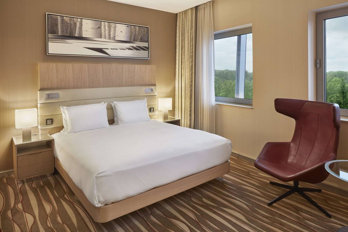 A hotel room with a bed, nightstands, a red chair, a window with a view, and a painting on the wall.