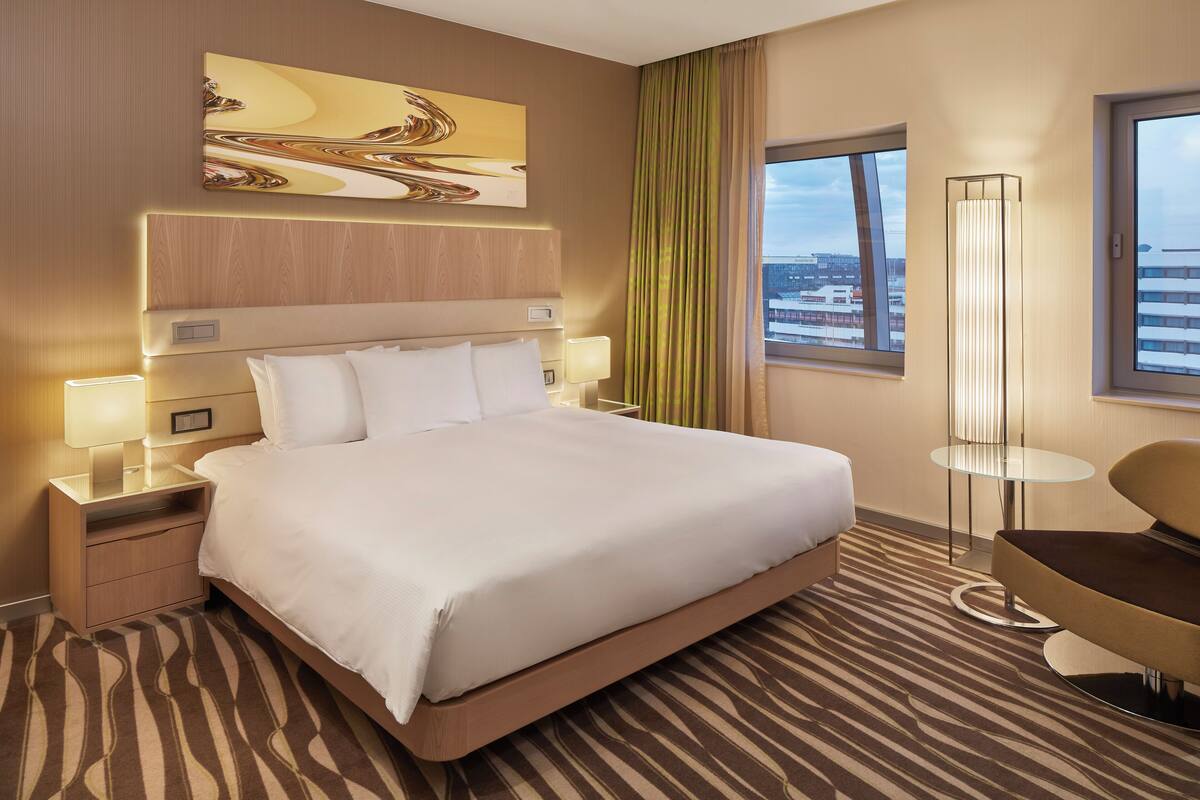 A hotel room with a large bed, bedside tables with lamps, a floor lamp, a chair, a view of the city through a window, and a painting on the wall.