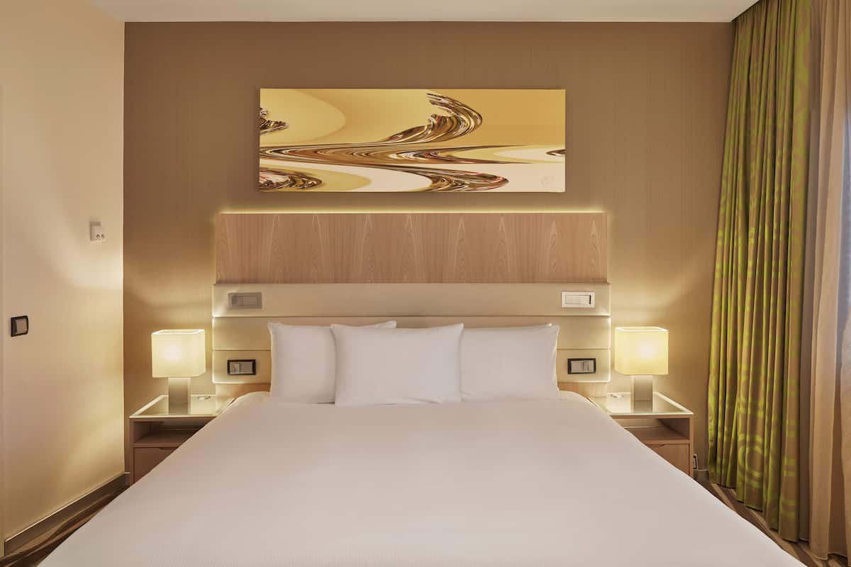 A hotel room with a large bed, two bedside tables with lamps, a headboard, and a wall-mounted abstract painting.