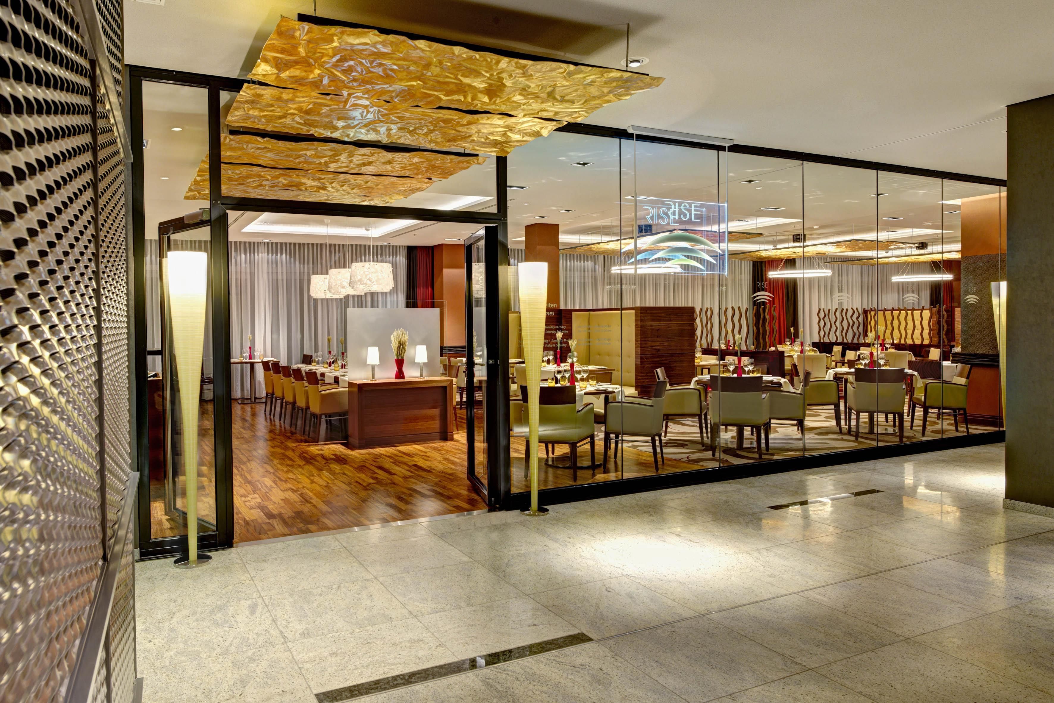 A modern hotel lobby with a glass partition, a reception desk, and a dining area with tables and chairs.