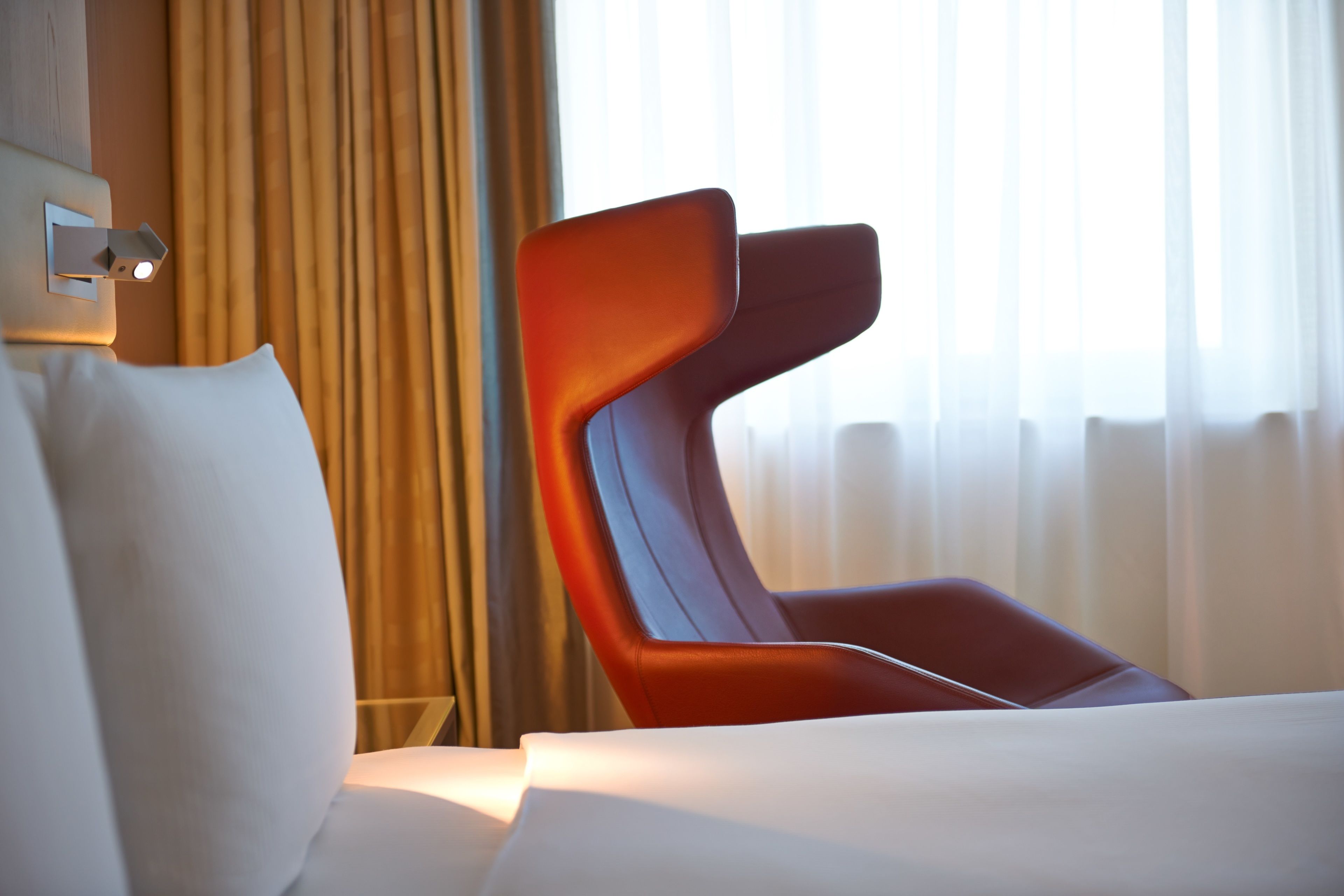 Una habitación de hotel moderna con una cama grande, una silla roja y un ventanal con cortinas.