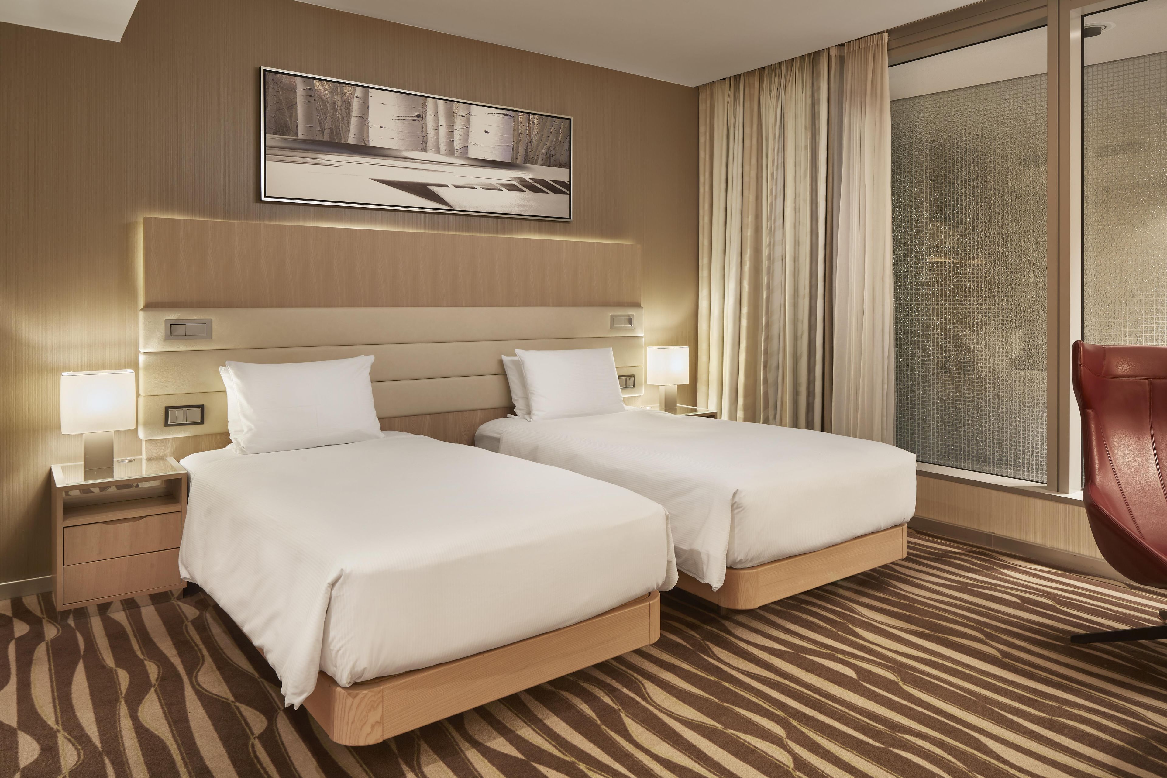 A hotel room with two beds, a large window with curtains, a wall-mounted TV, and a patterned carpet.