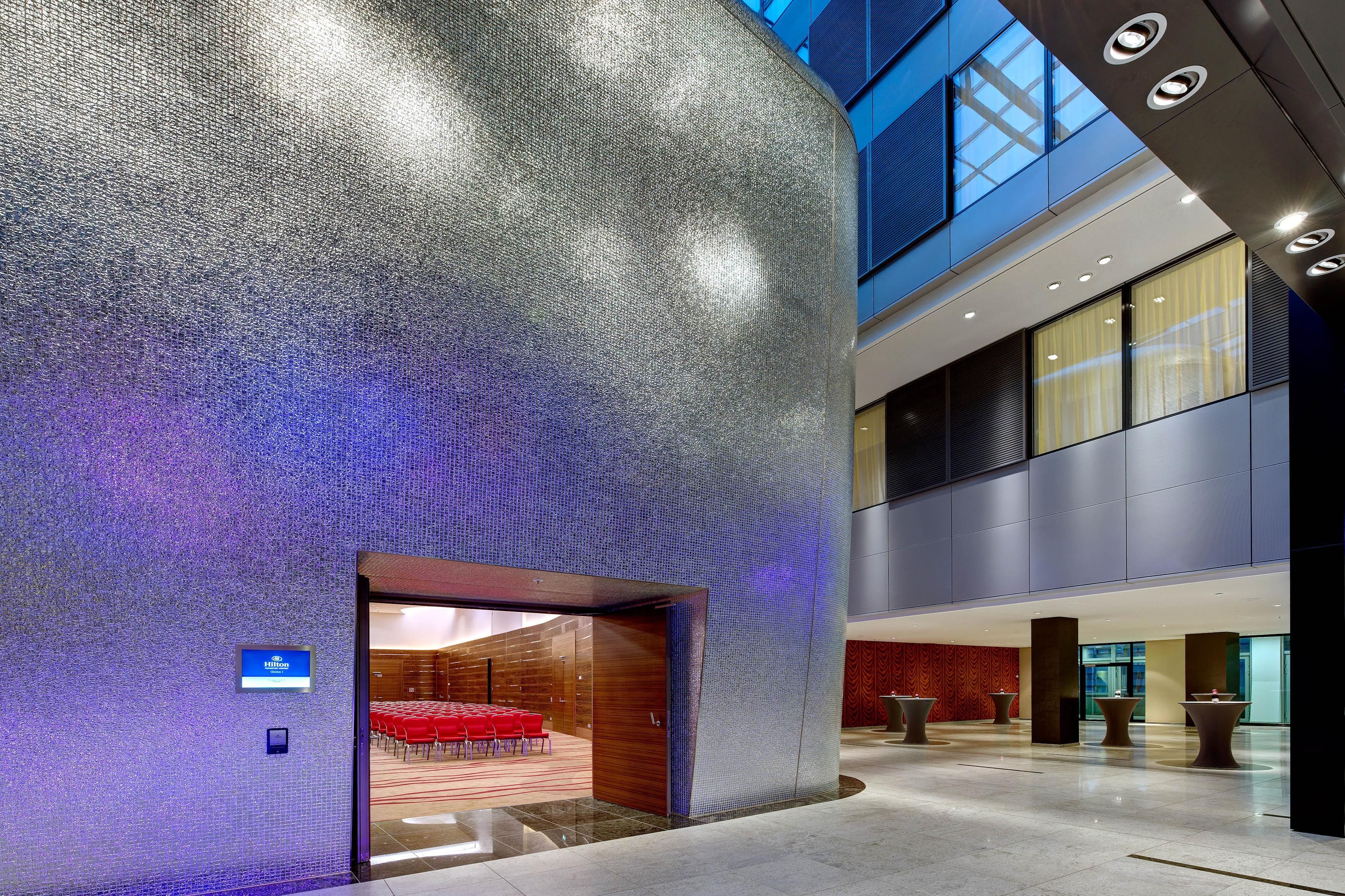 A modern hotel lobby with a textured wall, a seating area with red chairs, and a large entrance.
