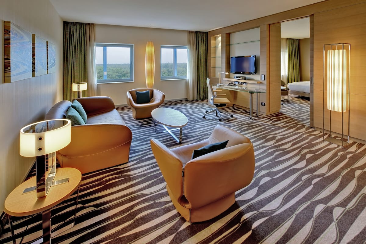 A hotel room with a sofa, armchair, desk, TV, and large windows.