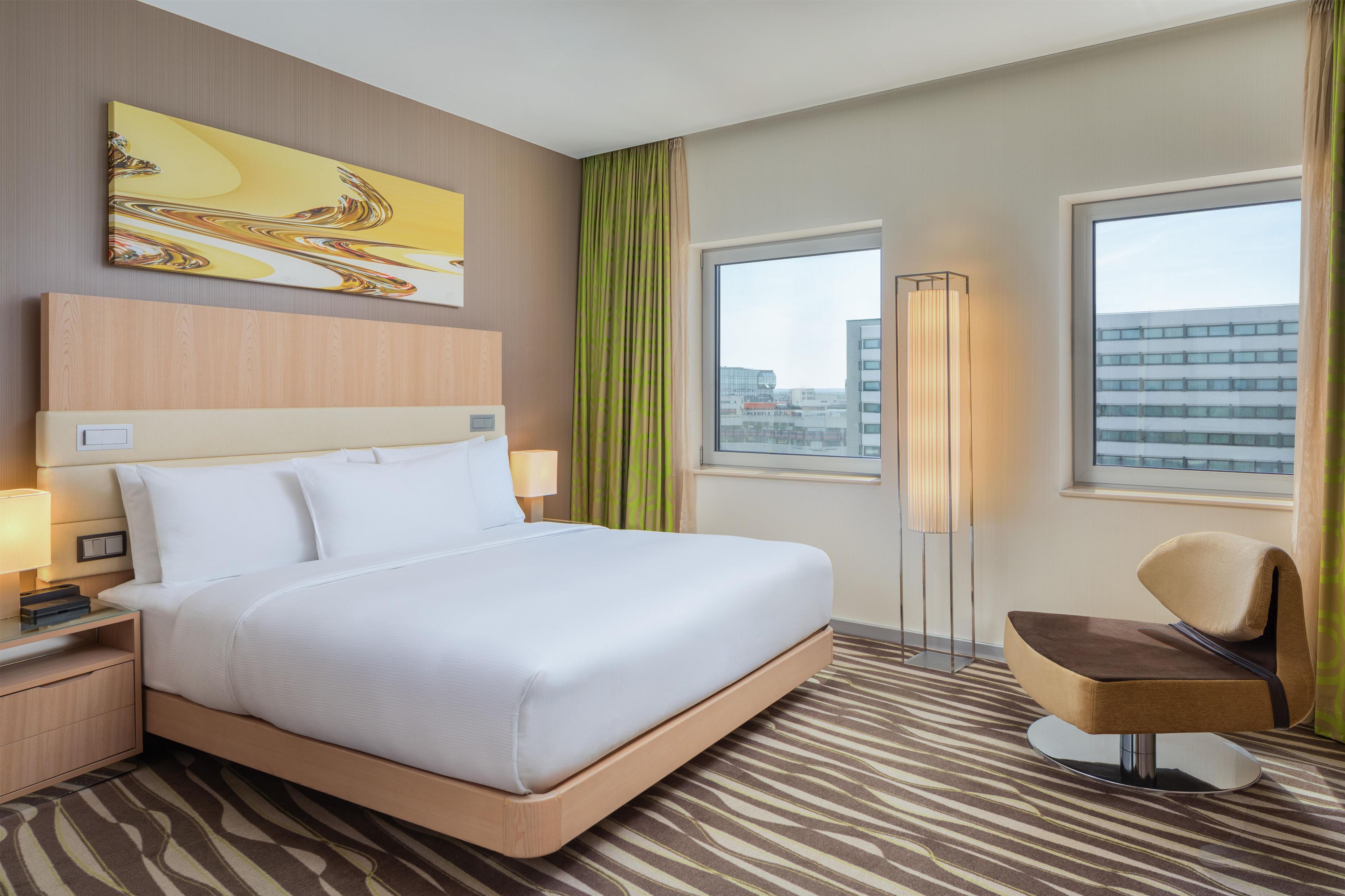 Habitación de hotel con una cama grande, una mesita de noche con lámpara, una silla, una lámpara de pie, una ventana con vista a edificios y un cuadro en la pared.