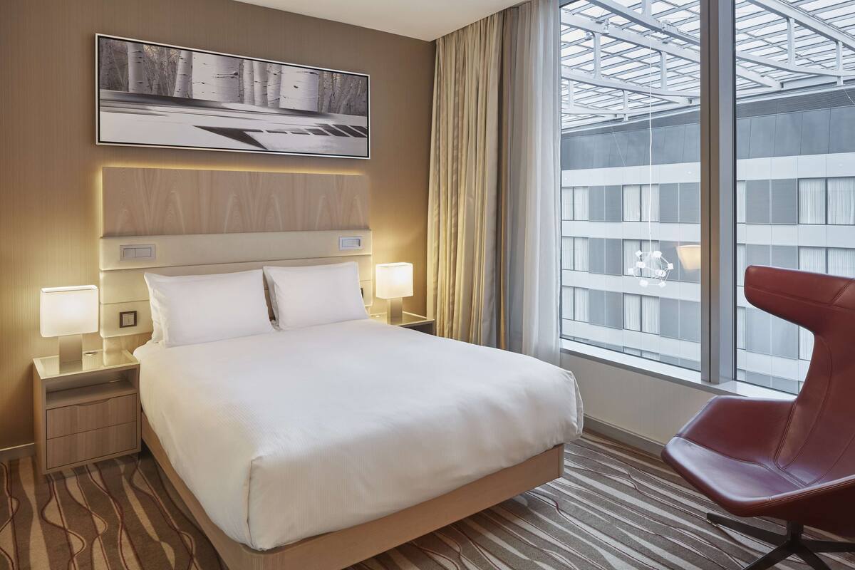 A hotel room with a large bed, two bedside tables with lamps, a red chair, and a large window with a view of buildings.