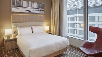 A hotel room with a large bed, two bedside tables with lamps, a red chair, and a large window with a view of buildings.