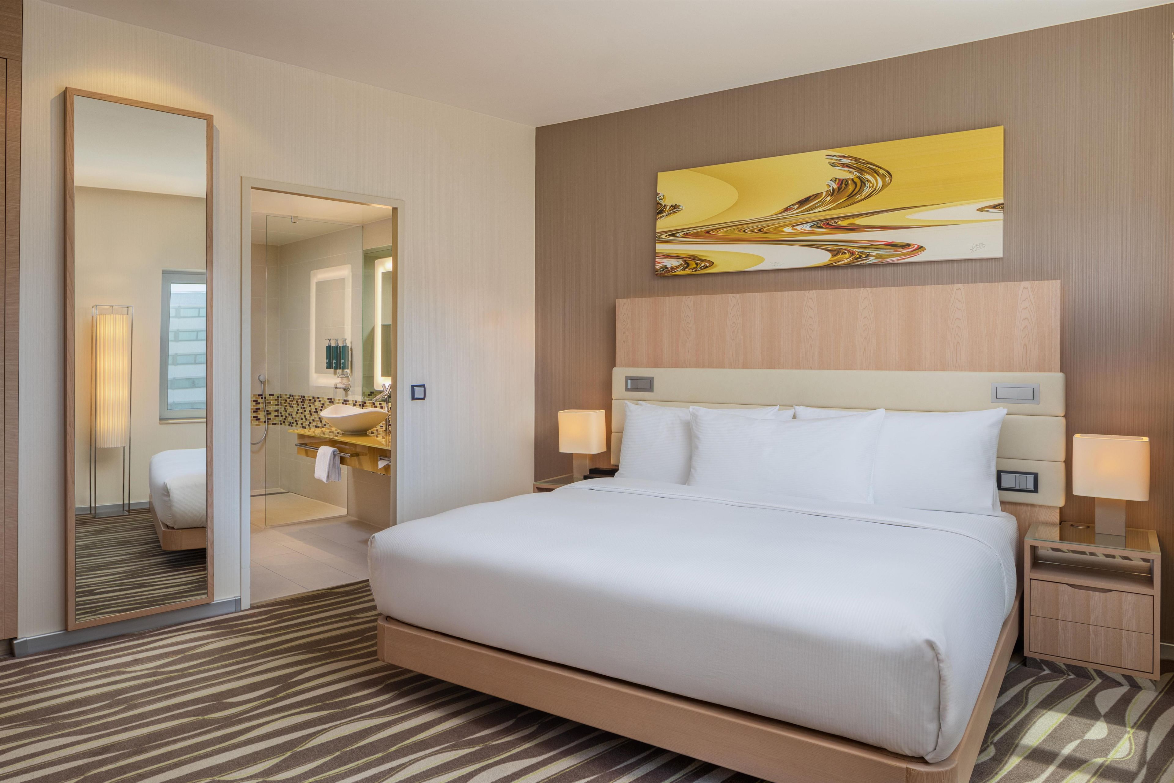 Ein Hotelzimmer mit einem großen Bett, einem Nachttisch mit Lampe, einem Spiegel und einem Wandbild.