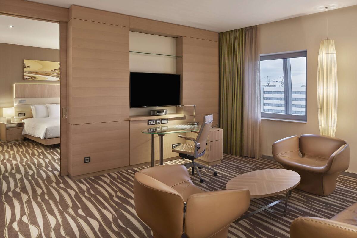 A hotel room with a bed, a desk with a flat-screen TV, a chair, a sofa, and a large window with curtains.