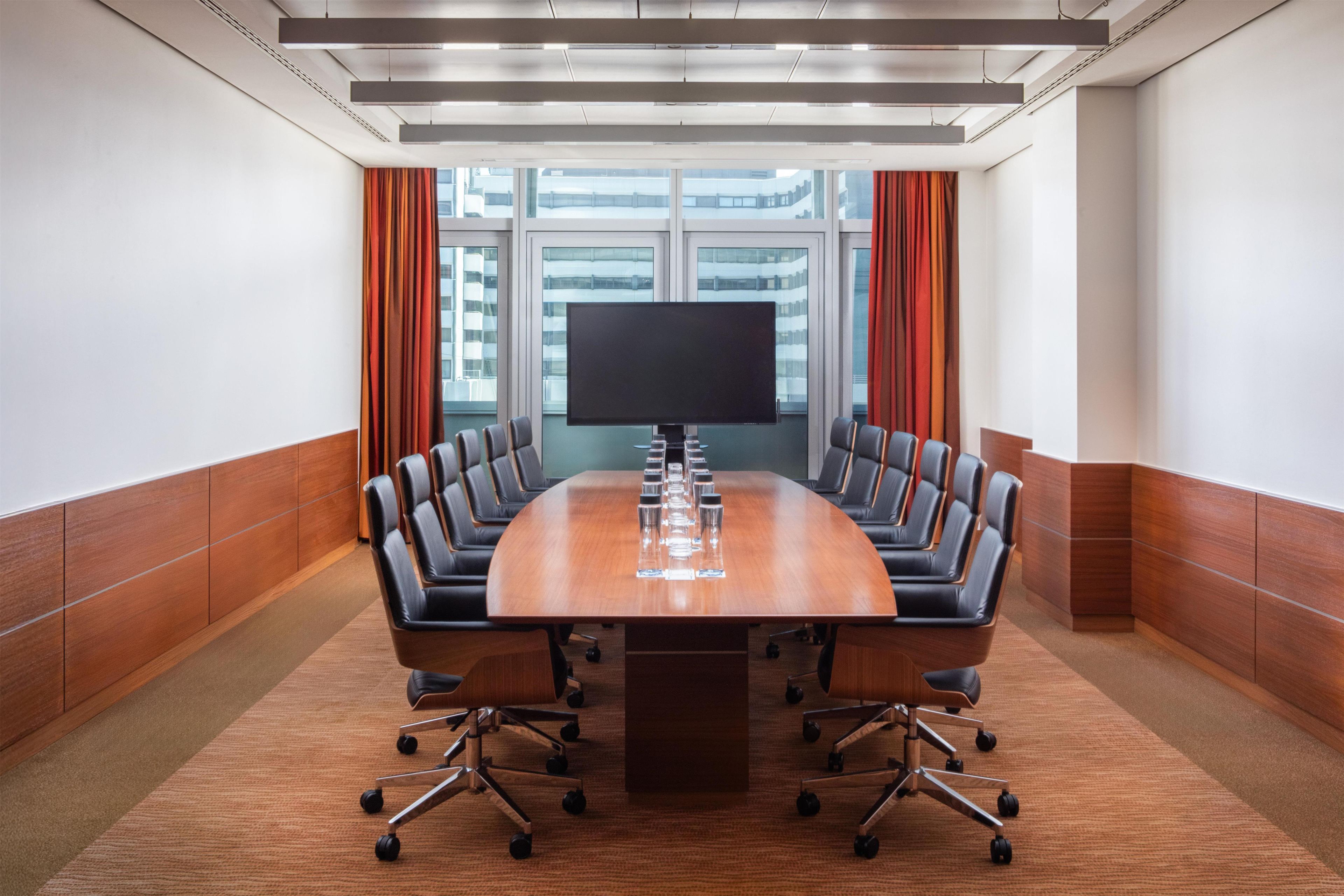 A conference room with a long table, multiple chairs, a large screen, and a view of buildings through large windows.