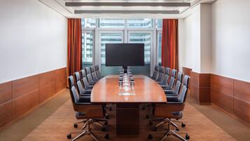 A conference room with a long table, multiple chairs, a large screen, and a view of buildings through large windows.