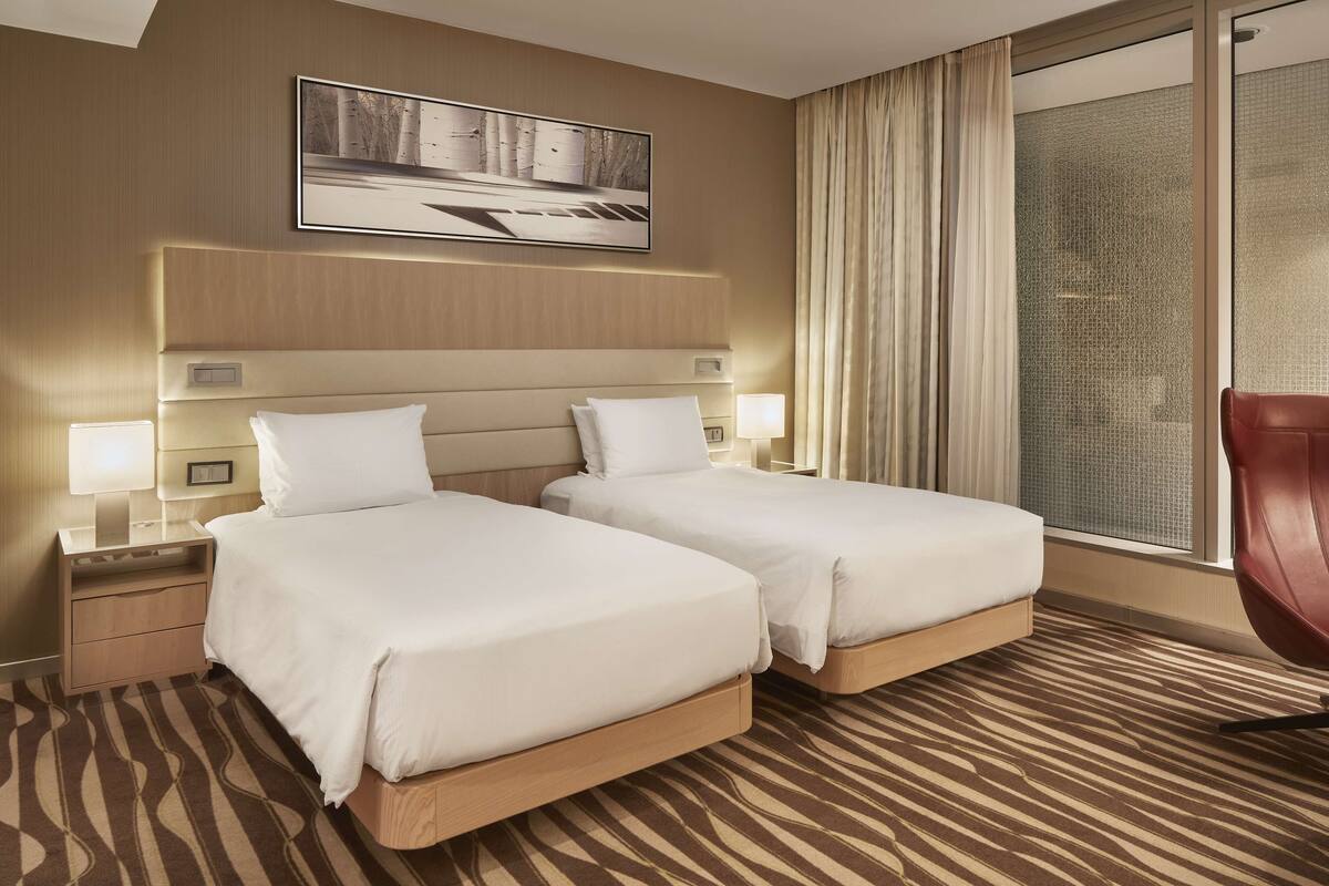 A hotel room with two beds, a large window with curtains, a red chair, and a framed picture on the wall.