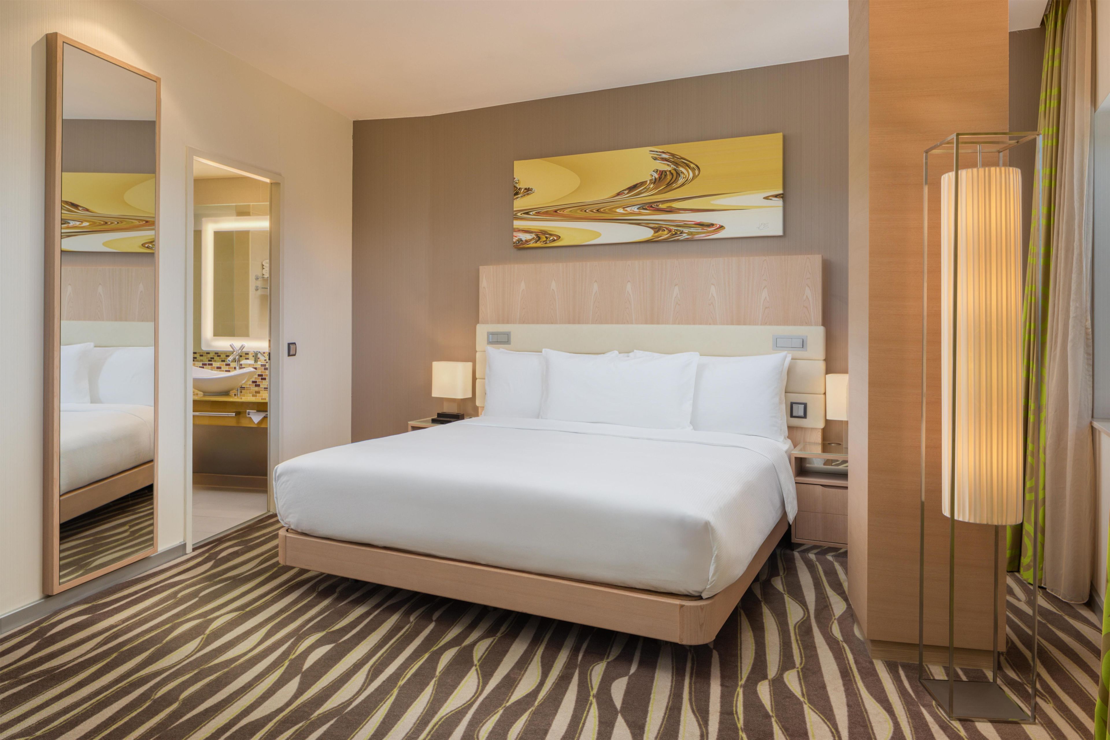 Una habitación de hotel con una cama grande, una mesita de noche con lámpara, un espejo y una obra de arte colgada en la pared.