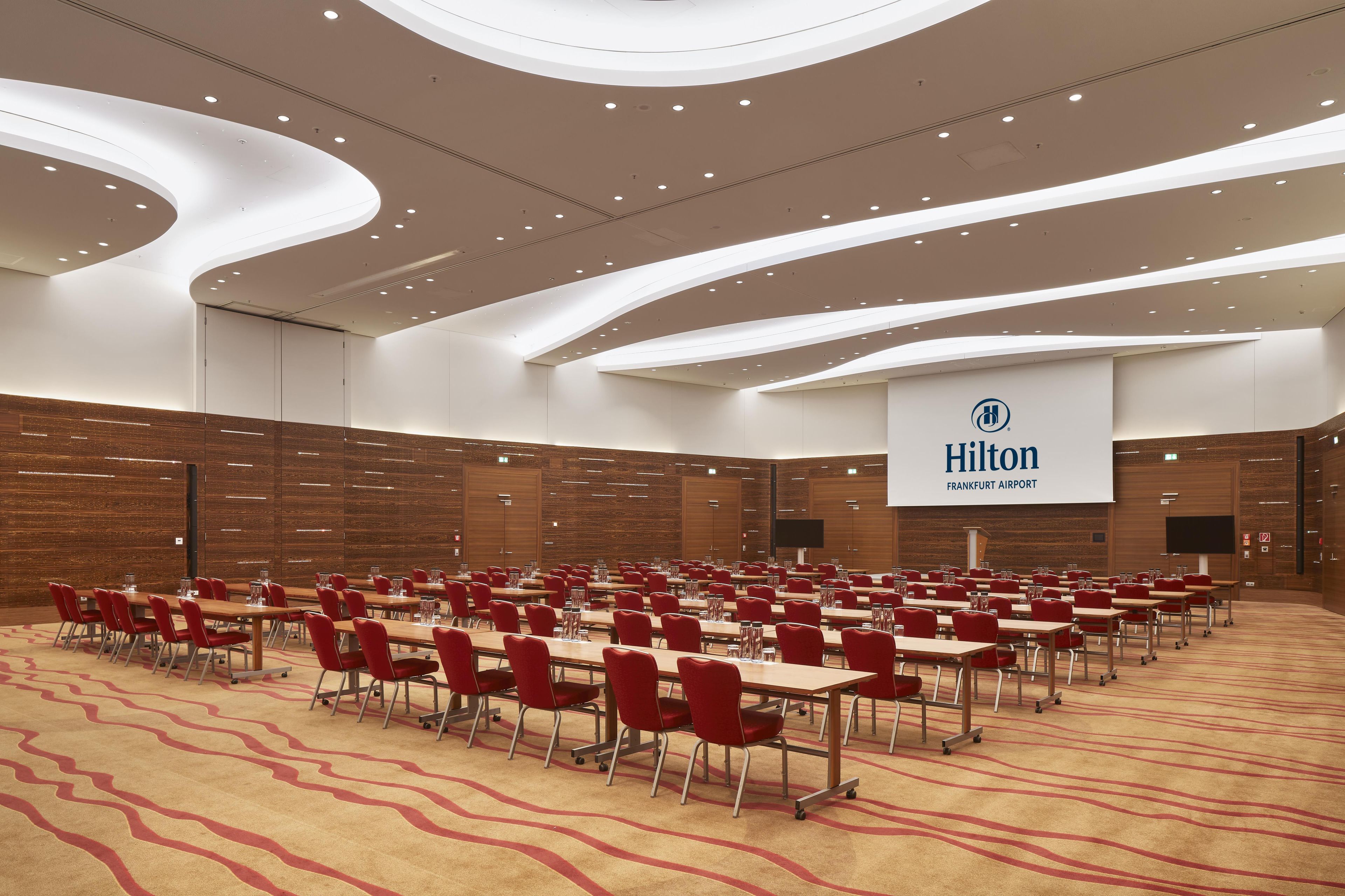 A large conference room with rows of tables and chairs, a modern ceiling design, and a sign for Hilton Frankfurt Airport.