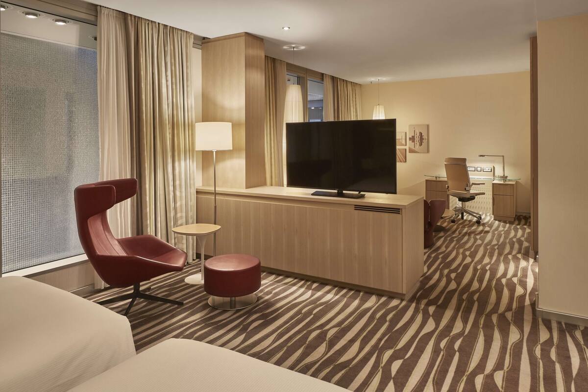 A modern hotel room with a large flat-screen TV, a red chair, a small round table, and a patterned carpet.