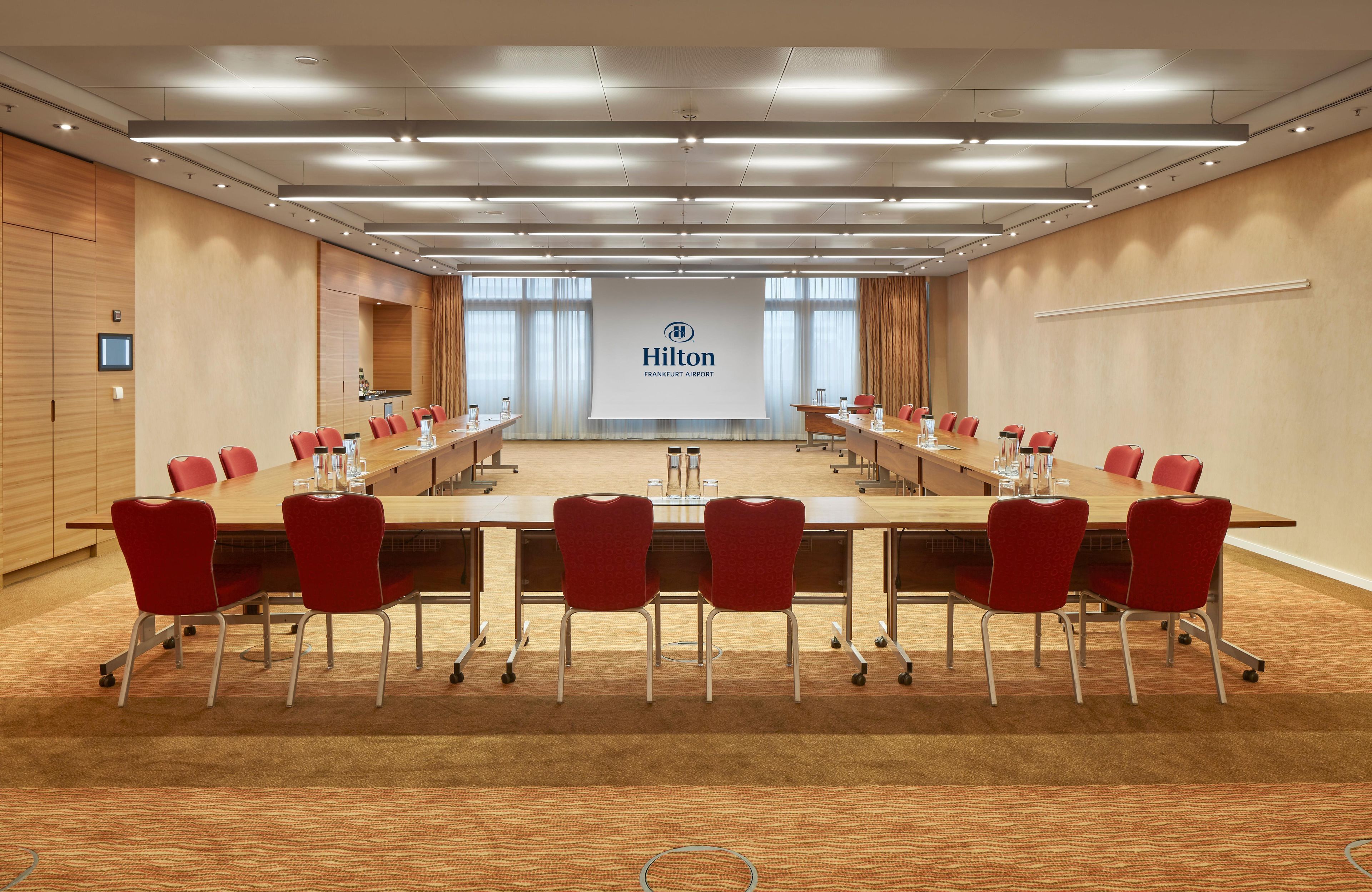 A conference room with a long table, red chairs, and a large screen displaying the Hilton Frankfurt Airport logo.