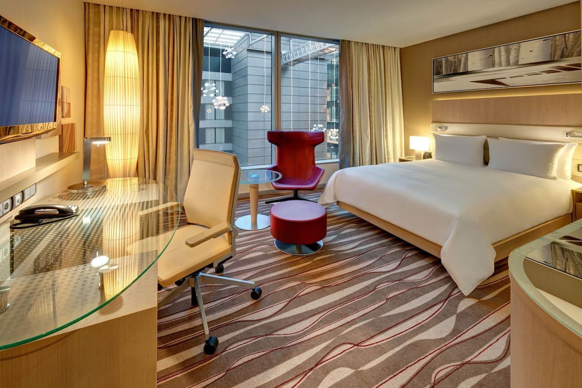 A hotel room with a bed, a desk with a telephone, a red chair, and a glass table.