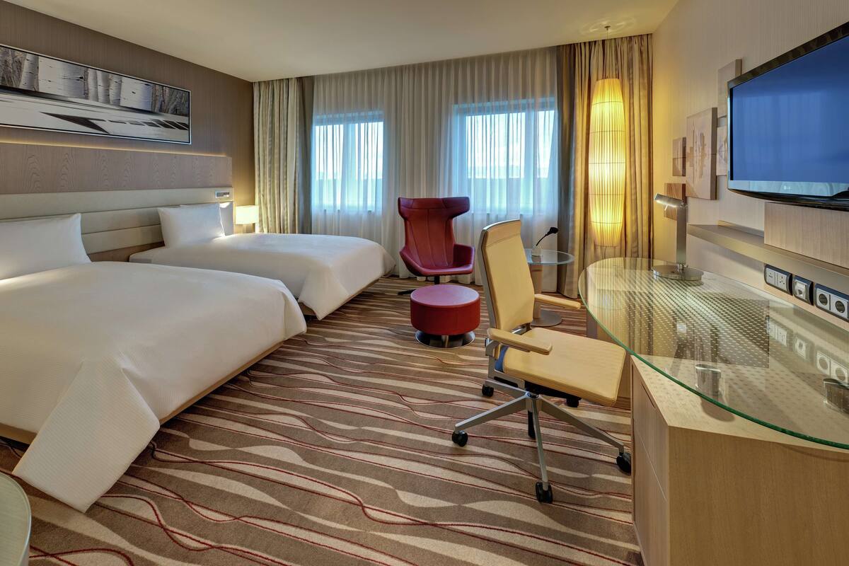 A hotel room with two beds, a red chair, a glass desk, and a TV.