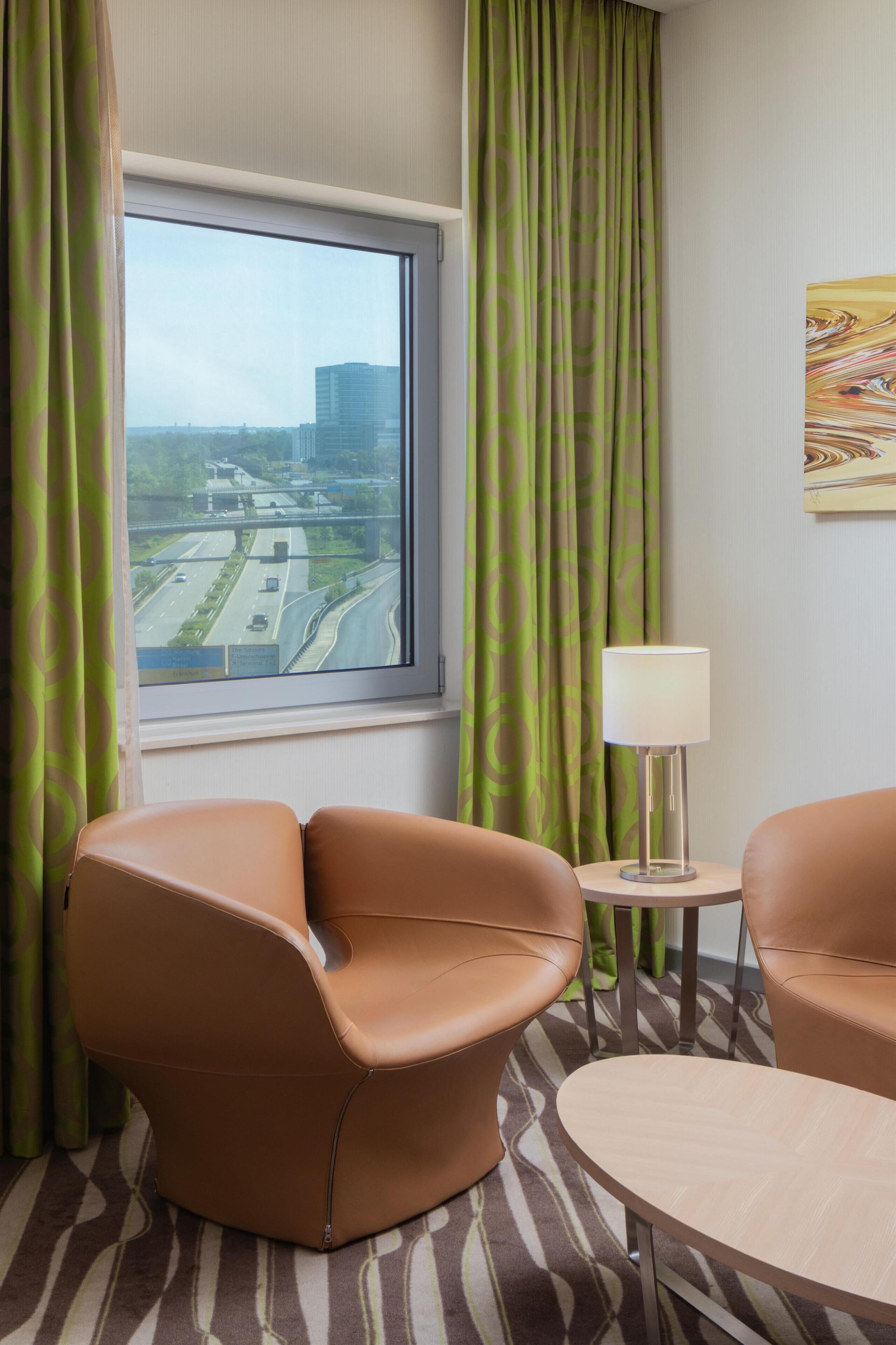 A hotel room with a large window, green curtains, a brown leather chair, a small round table, and a lamp.