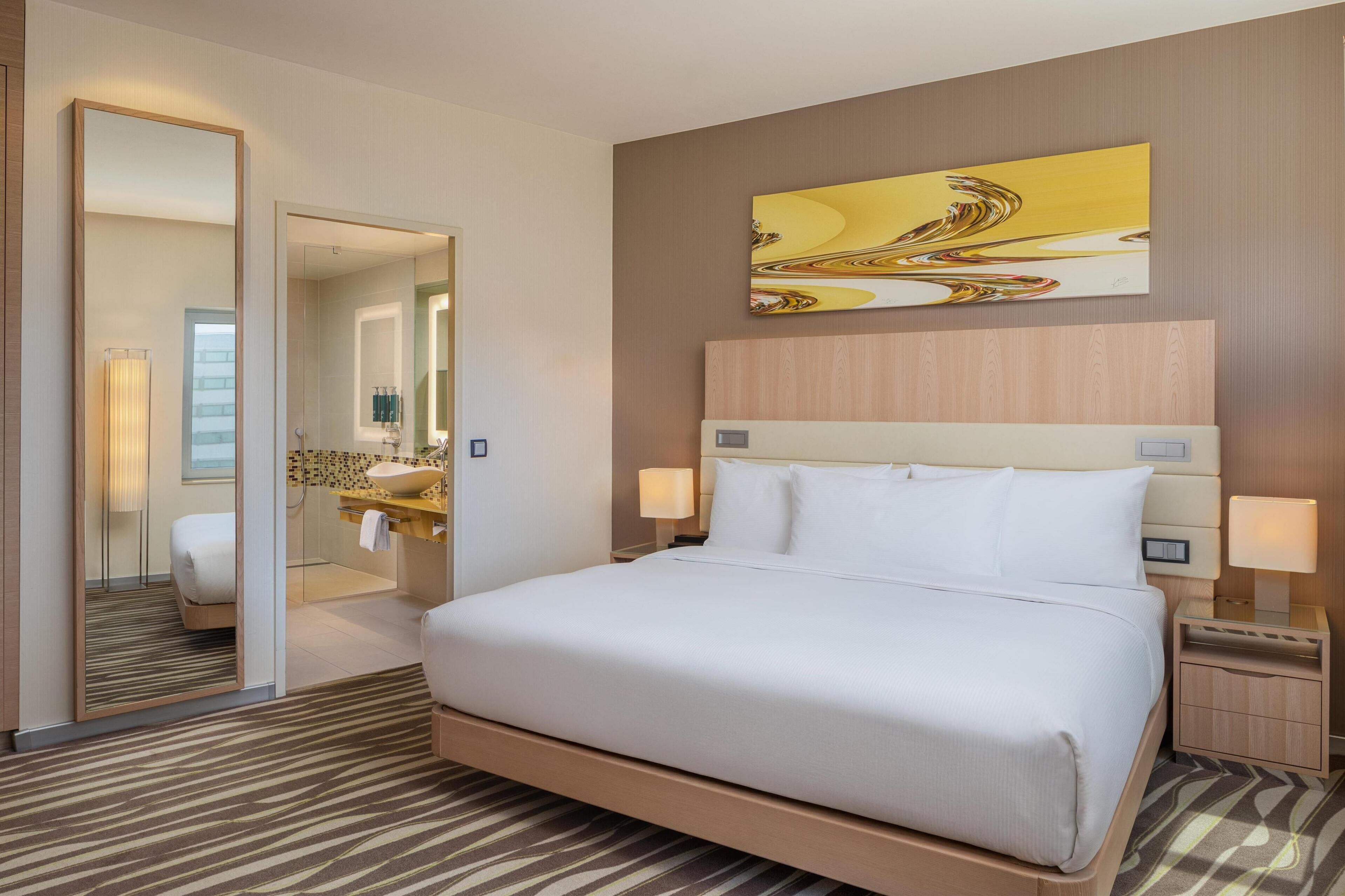 Habitación de hotel con una cama grande, una mesita de noche con lámpara, un espejo y un baño visible a través de una puerta abierta.