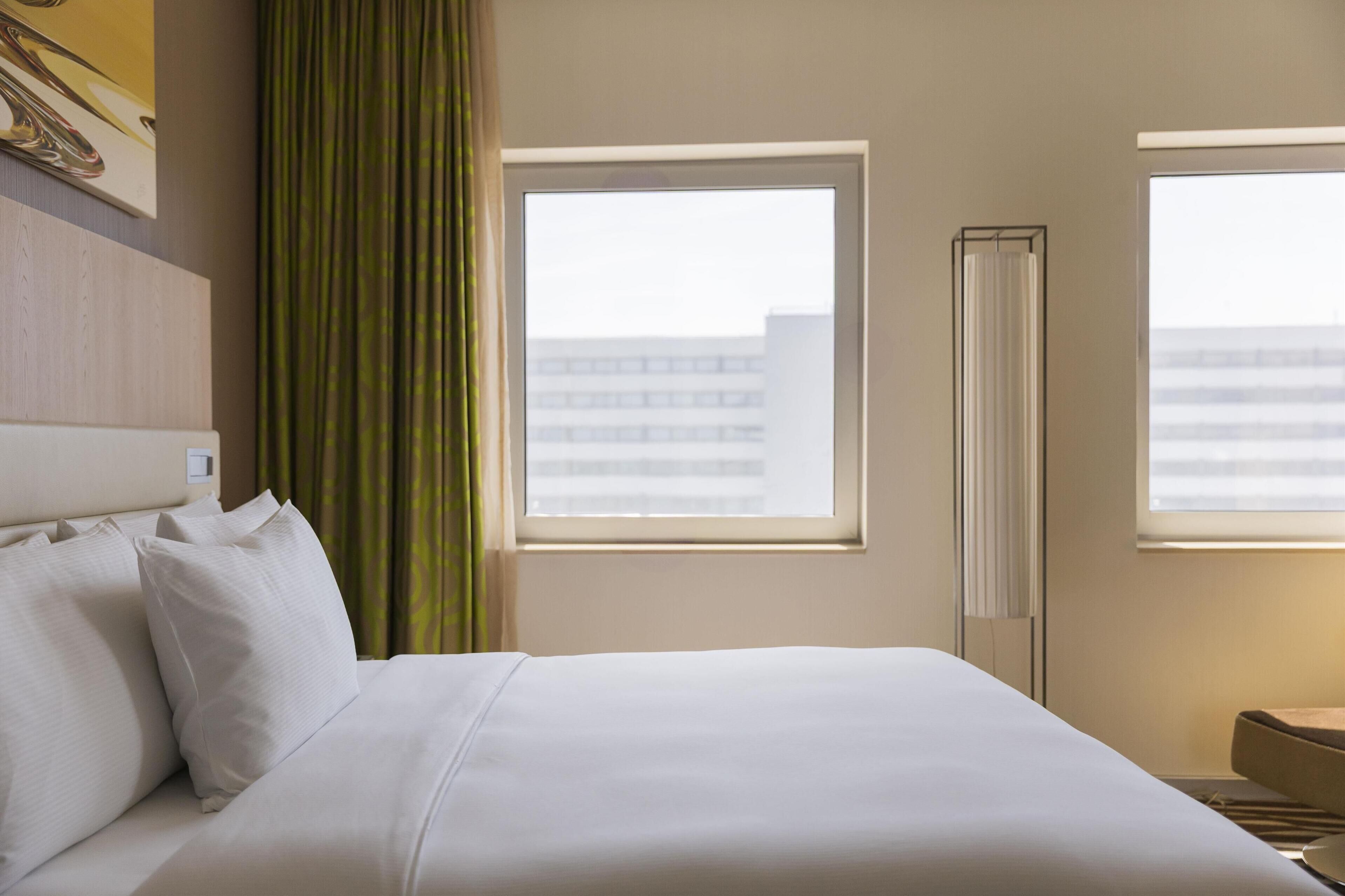 Una habitación de hotel con una cama grande, dos ventanas, un radiador y vistas a un edificio.