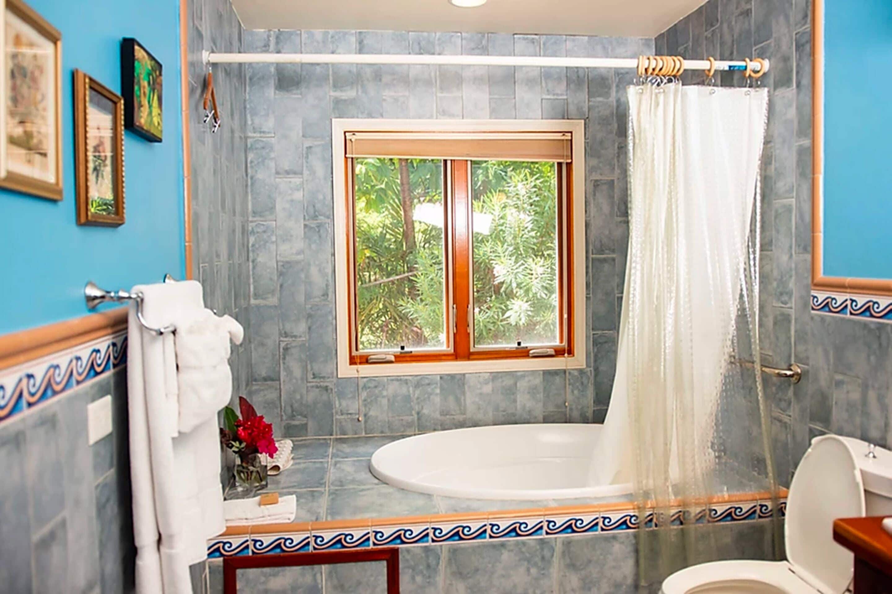 A bathroom with a bathtub, a toilet, a window with a view of greenery, and decorative tiles.