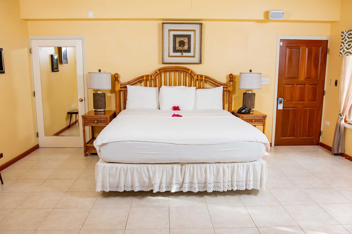 A neatly made bed with a white bedspread and a decorative skirt, two bedside tables with lamps, a wooden headboard, a framed picture on the wall, and a door with a mirror reflection.