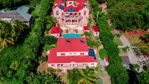 A large, multi-story house with a red roof and solar panels, surrounded by lush greenery and palm trees. - The Villas at Sunset Lane (St. John's)