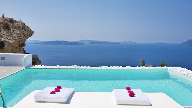 Une piscine bordée de chaises longues blanches et de serviettes, offrant une vue imprenable sur une mer d’un bleu limpide et une côte rocheuse.