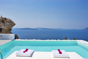 A pool with white lounge chairs and towels, overlooking a clear blue sea and rocky coastline. - Villaki Oia (Santorini)