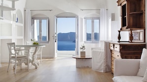 Une pièce spacieuse avec une grande porte ouverte donnant sur un balcon avec vue sur la mer. On y trouve une table à manger avec des chaises, un canapé blanc et un meuble en bois.