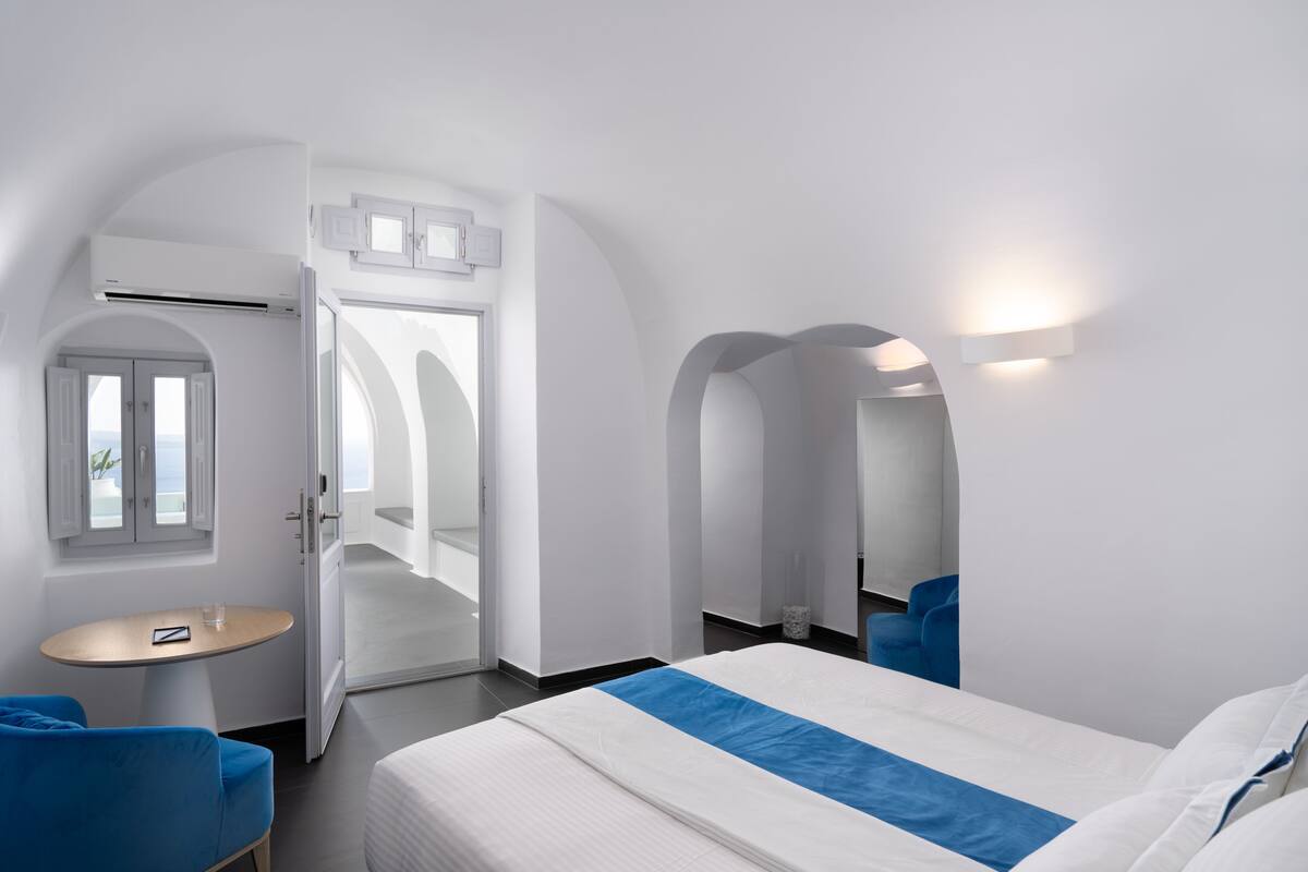 Ein modernes Hotelzimmer mit einem großen Bett, einem runden Tisch und blauen Stühlen. Eine Tür führt zu einem Balkon mit Aussicht.