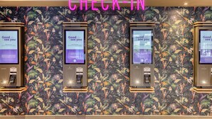 Un espace d’enregistrement agrémenté d’un papier peint à motifs floraux, de trois bornes d’enregistrement en libre-service et d’un panneau néon indiquant « ENREGISTREMENT ».