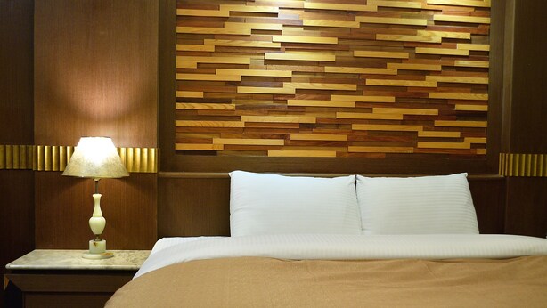 Een net opgemaakt bed met witte lakens en een beige plaid, een houten hoofdbord met een geometrisch patroon, en een nachtkastje met een lamp.