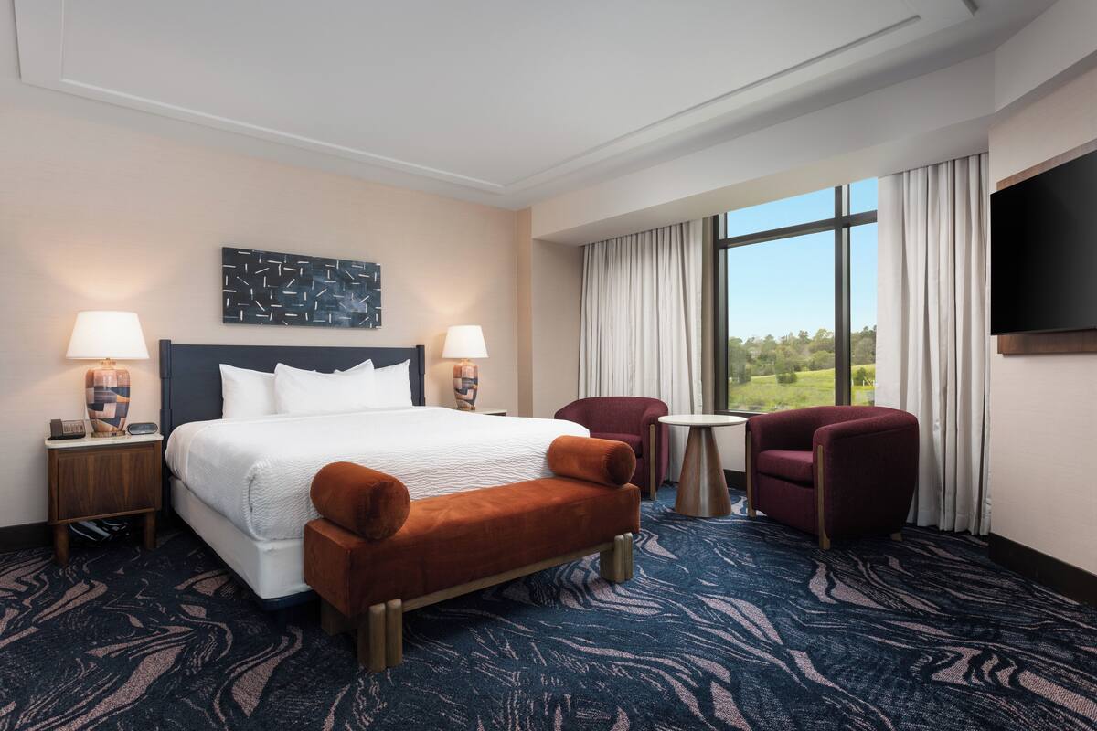 A hotel room with a large bed, two side tables with lamps, a small round table, two armchairs, a flat-screen TV, and a large window with a view of greenery.