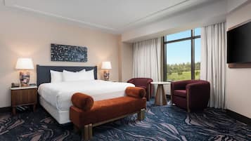 A hotel room with a large bed, two side tables with lamps, a small round table, two armchairs, a flat-screen TV, and a large window with a view of greenery.