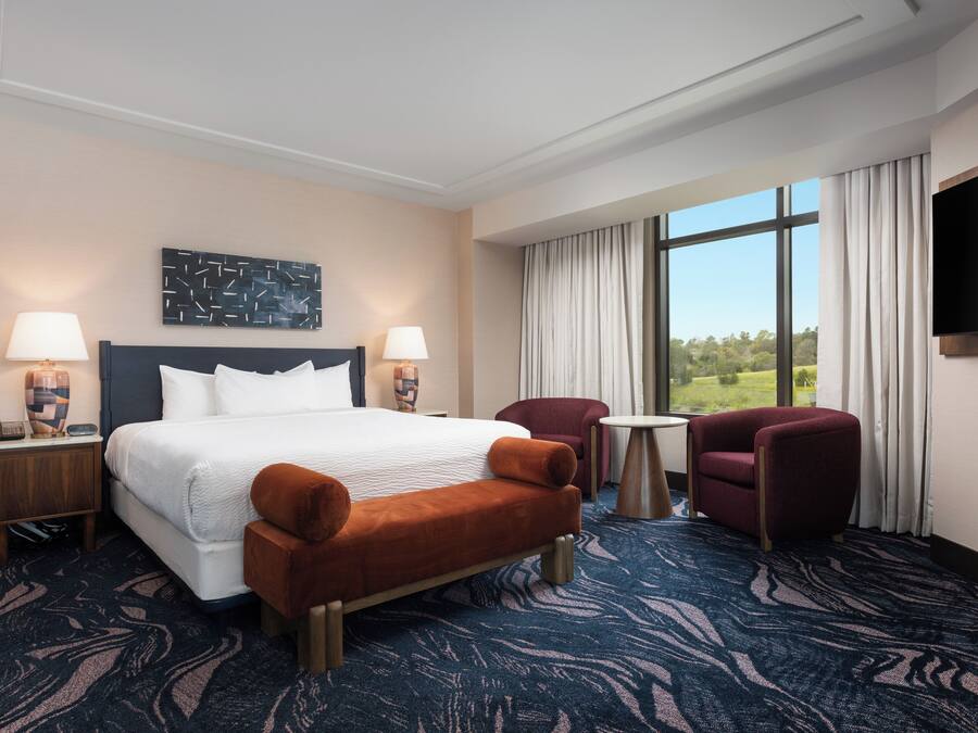 A hotel room with a large bed, two side tables with lamps, a small round table, two armchairs, a flat-screen TV, and a large window with a view of greenery.