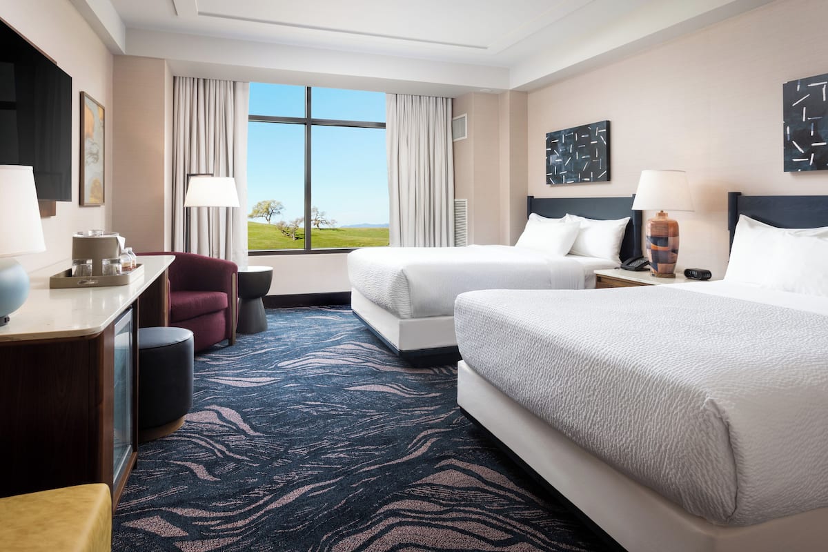 A hotel room with two beds, a desk with a lamp, a chair, and a view of the outdoors.