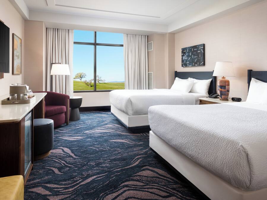 Habitación de hotel con dos camas, un escritorio con lámpara, una silla y vistas al exterior.