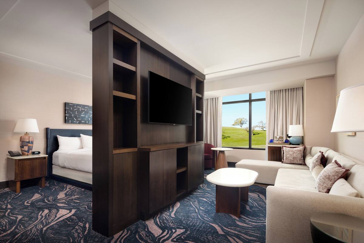 A hotel room with a large flat-screen TV, a sofa, a bed, a desk, and a window with a view of the outdoors.
