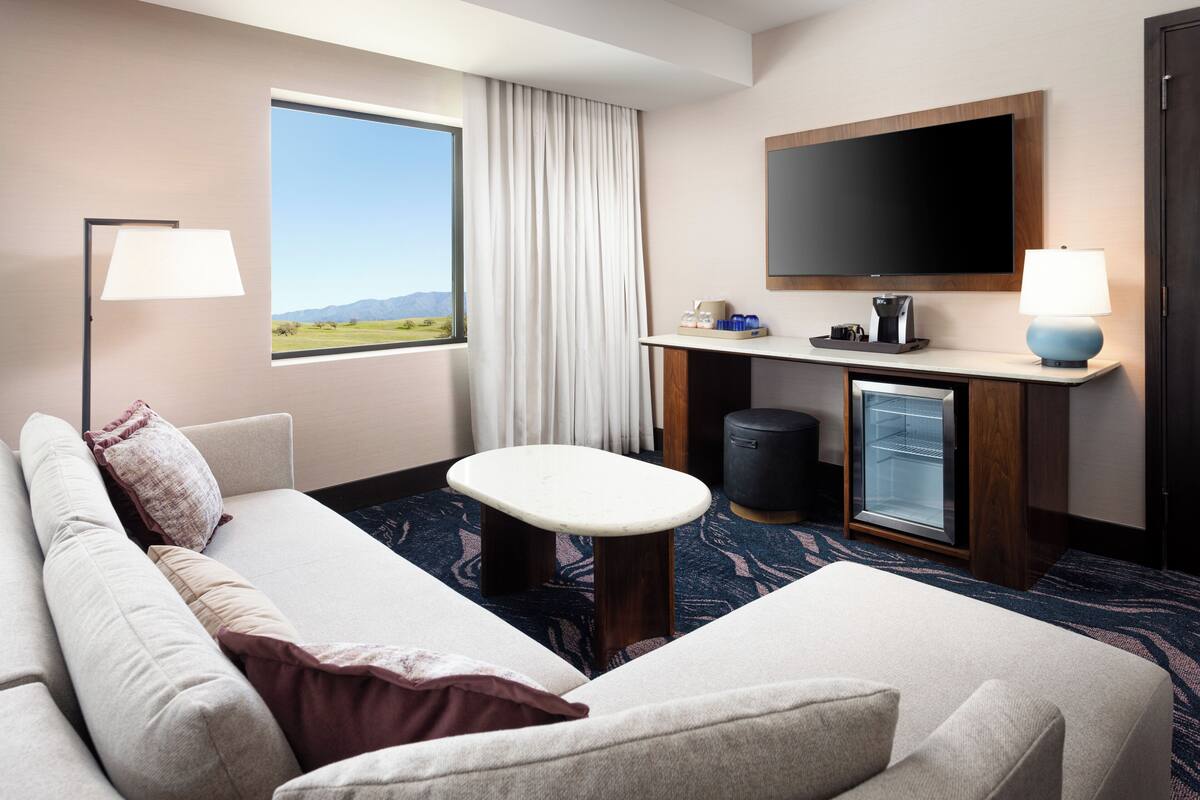 A hotel room with a sofa, a small table, a TV, a minibar, and a window with a view of the outdoors.
