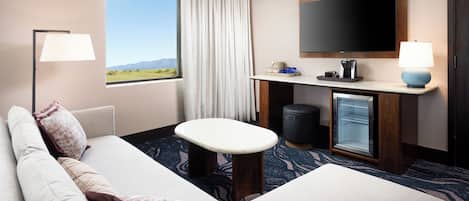 Habitación de hotel con sofá, una mesita, televisión, minibar y ventana con vistas al exterior.