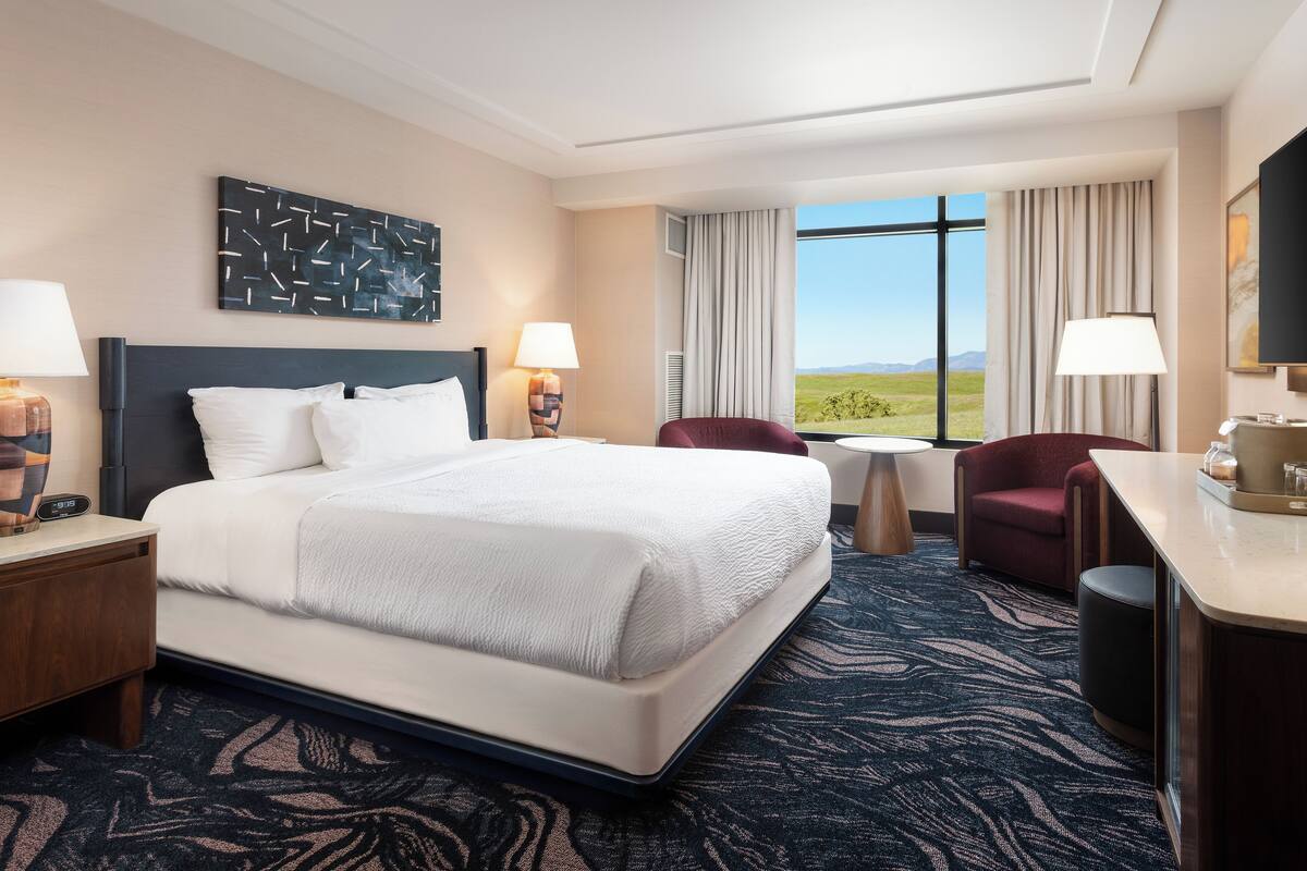 A hotel room with a large bed, two bedside tables, a desk, a chair, and a large window offering a view of the outdoors.