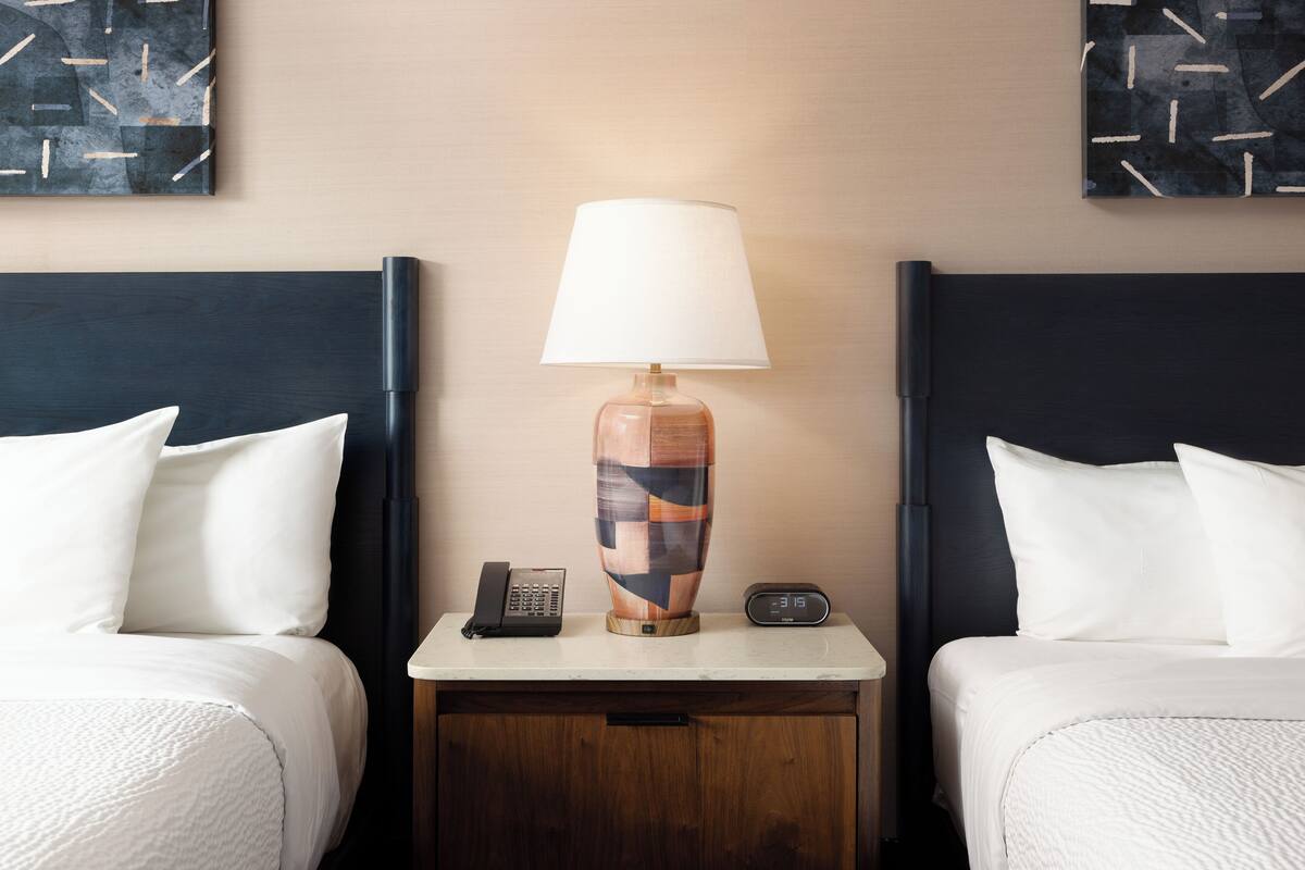 A hotel room with two beds, a nightstand with a lamp, a phone, and a clock.
