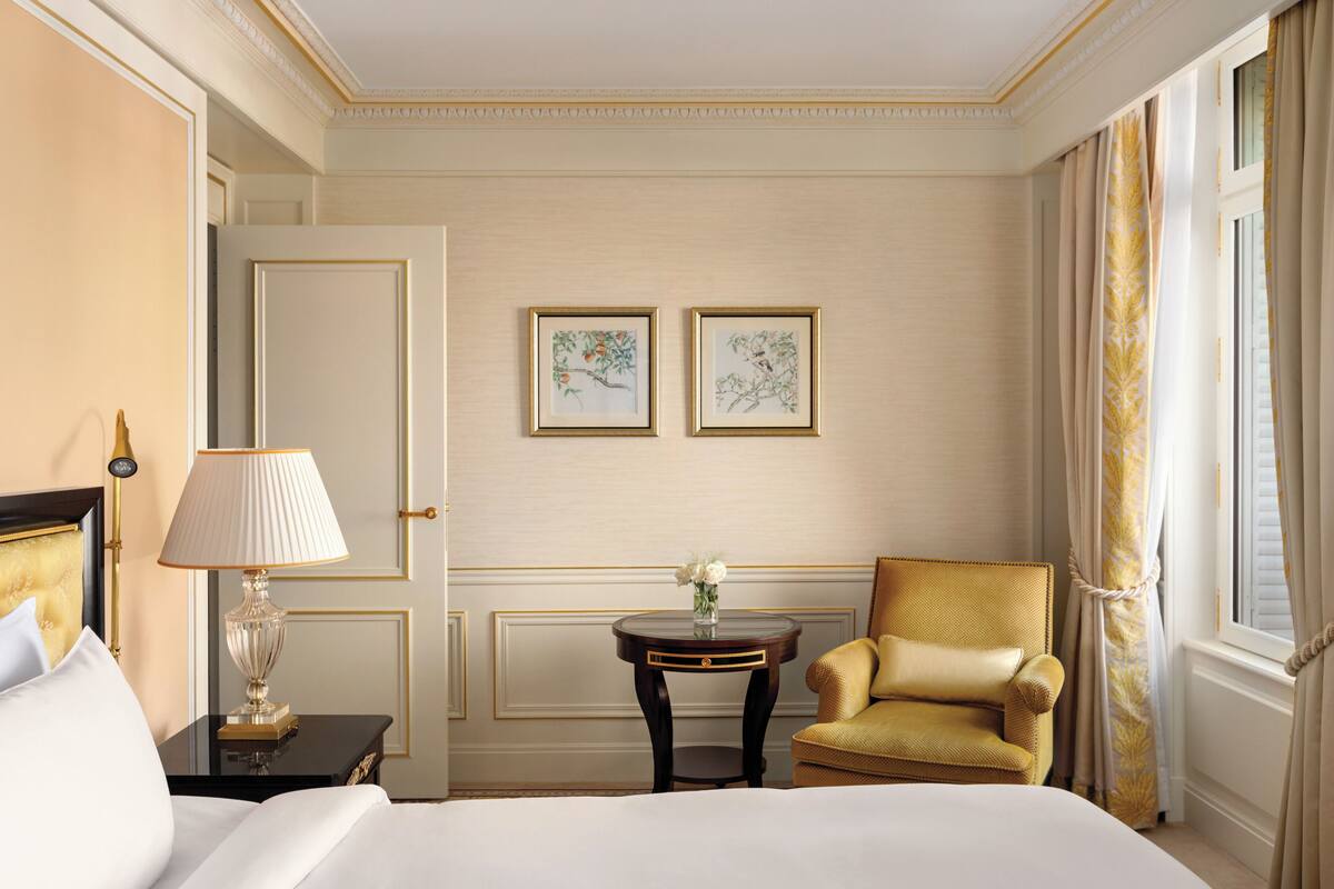 Une chambre d’hôtel avec un lit, une table de chevet avec une lampe, une petite table sur laquelle est posé un vase de fleurs, un fauteuil doré et deux reproductions botaniques encadrées accrochées au mur.